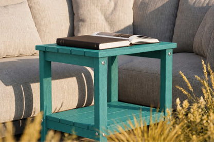 A teal outdoor side table with a slatted design, styled next to a gray fabric sofa with books on top.
