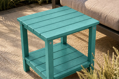 A teal outdoor side table with a two-tier design, shown next to a patio sofa on a stone patio.