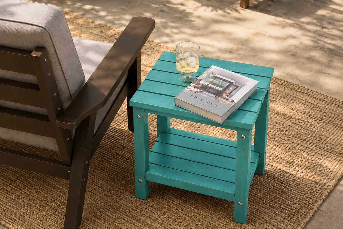 A turquoise outdoor side table with two shelves, styled next to a dark brown patio armchair on a woven jute rug. A glass of white wine and a book are placed on the table.