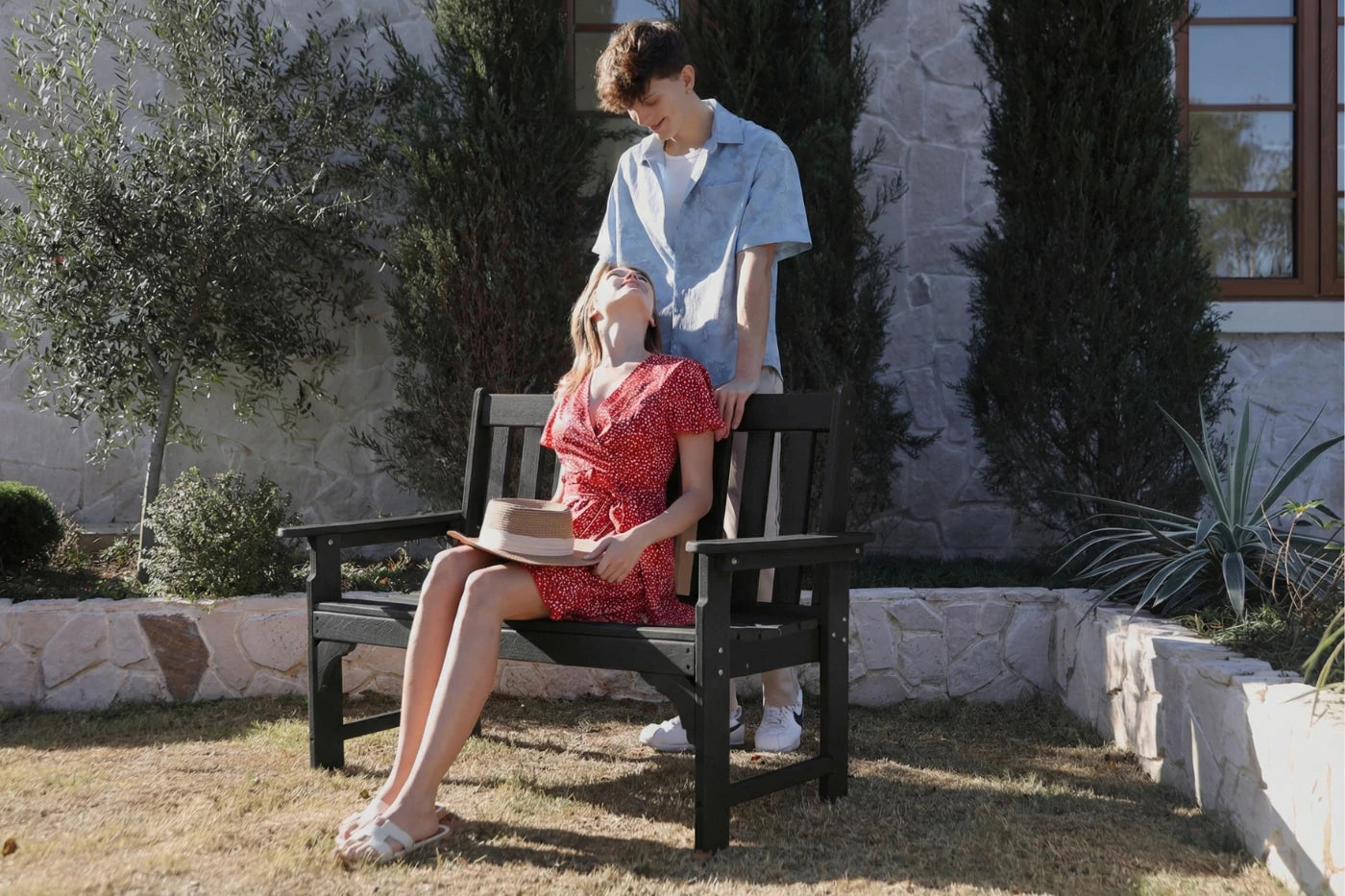 Two people sitting on a bench outdoors with plants and a building in the background