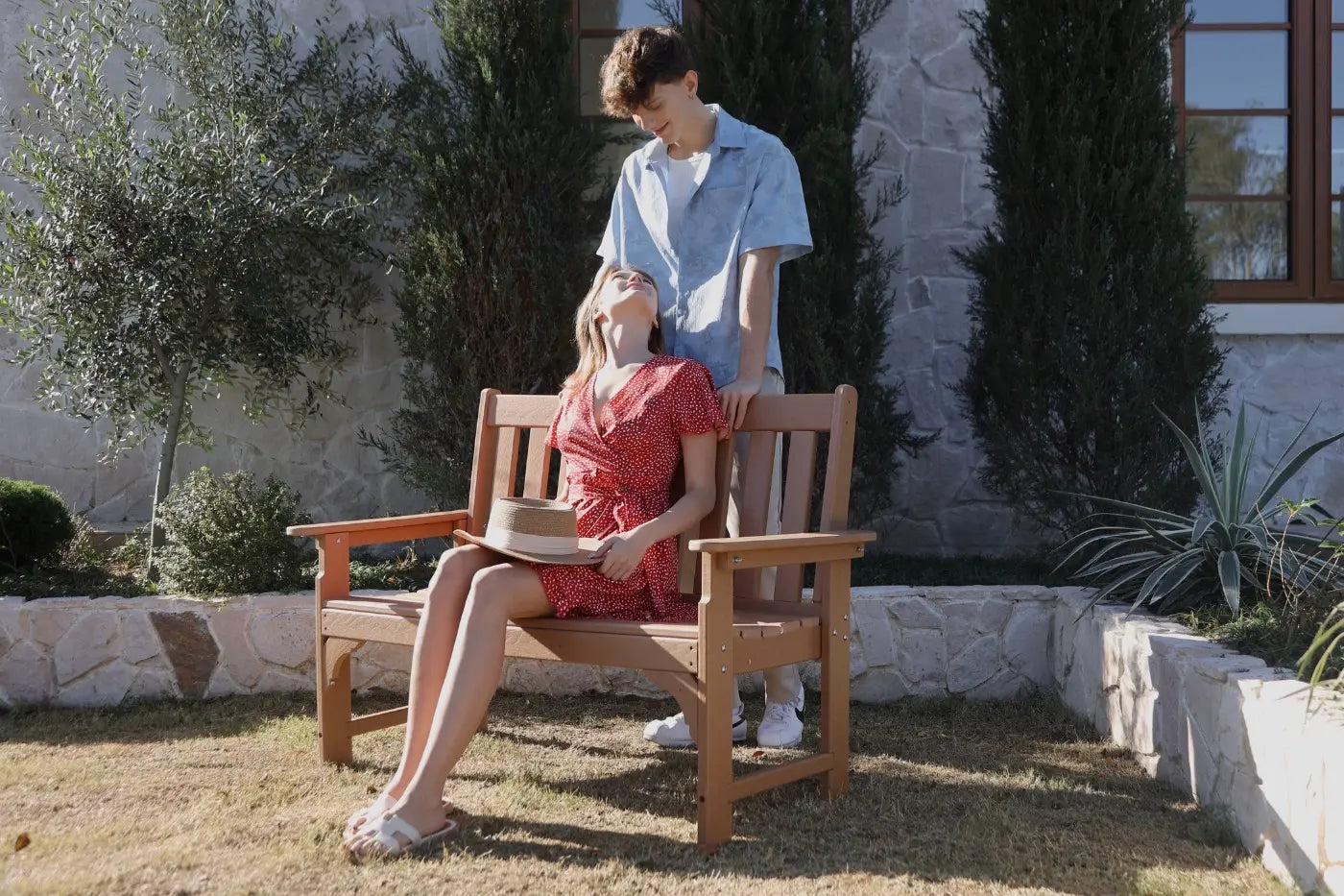 Two people sitting on a wooden bench outdoors with a stone building and trees in the background.
