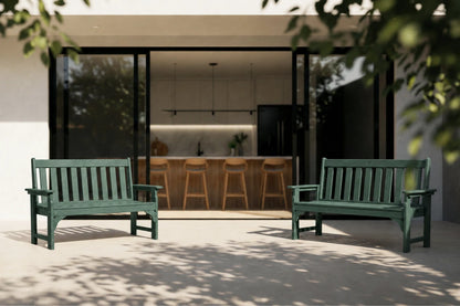 Two dark green outdoor bench chairs placed on a patio in front of a modern open-tile house with a kitchen visible inside.