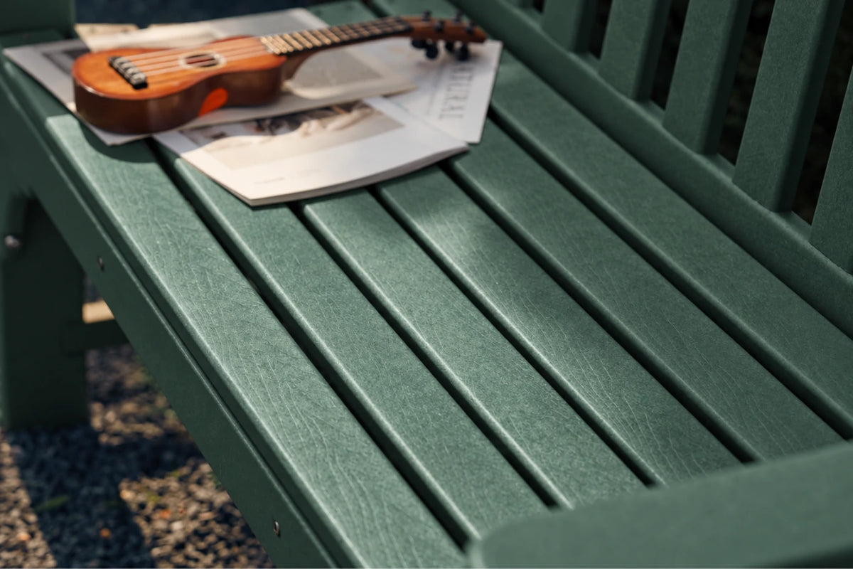 A close-up of a dark green slatted outdoor bench with a small brown ukulele resting on top of a newspaper, with gravel visible underneath.