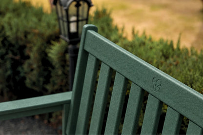 A close-up of a dark green slatted outdoor bench with a small heart-shaped logo engraved on the backrest, set against a blurred garden background with a black lamp post.
