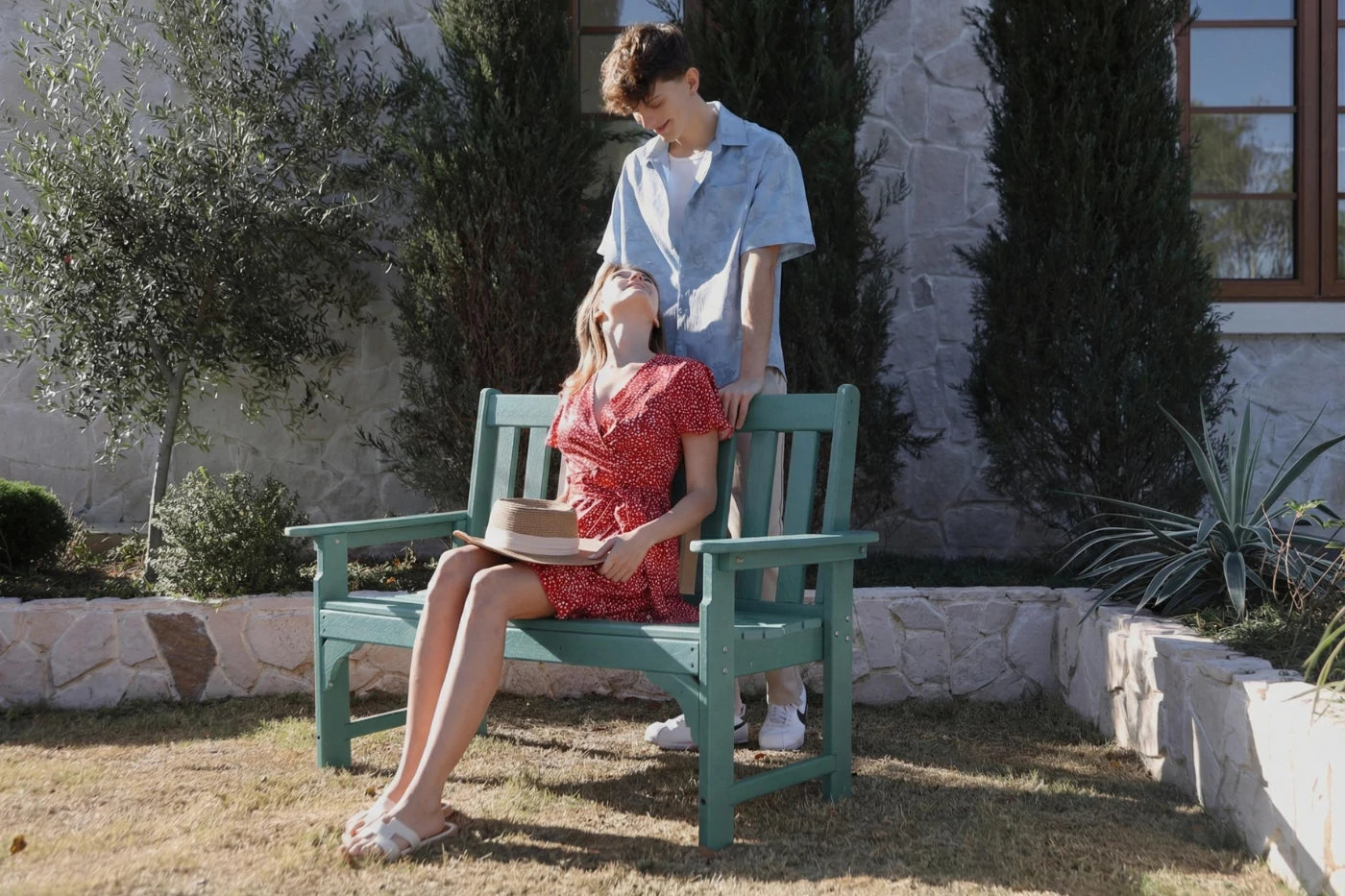 Two people sitting on a green bench outdoors with trees and a building in the background.