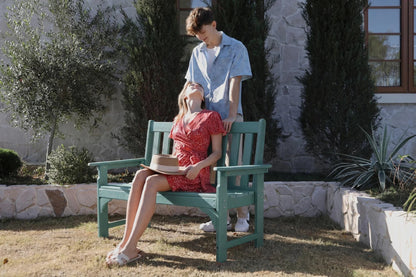 Two people sitting on a green bench outdoors with trees and a building in the background.