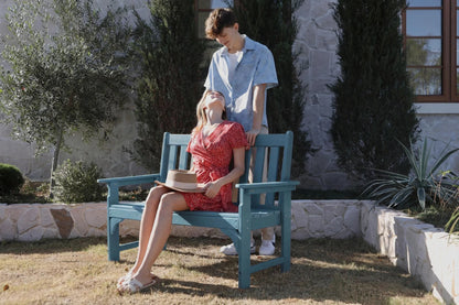 Two people sitting on a blue bench outdoors with a stone wall and trees in the background.
