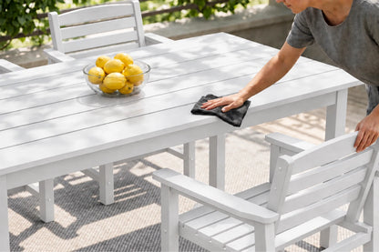 A white outdoor dining table and matching chairs are shown in a patio setting, with a person wiping the tabletop with a cloth and a bowl of lemons on the table.