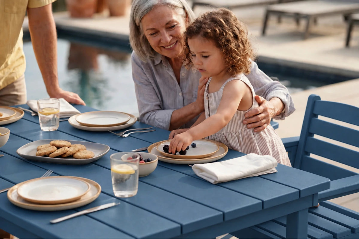 An outdoor dining scene by a pool where a woman and a young girl sit at a blue table, sharing food. The image features a woman in a light gray shirt and a young girl in a white dress, with another person partially visible on the left.