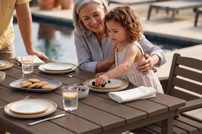 A grandmother and young daughter share a meal at an outdoor wooden dining table by a swimming pool. The table is set with plates, glasses, and a plate of cookies.
