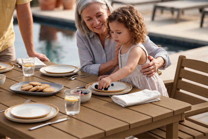 A woman and a young girl sit at an outdoor dining table by a pool, sharing a meal with plates, glasses, and cookies. The woman wears a light blue button-up shirt, and the girl wears a white sleeveless dress.