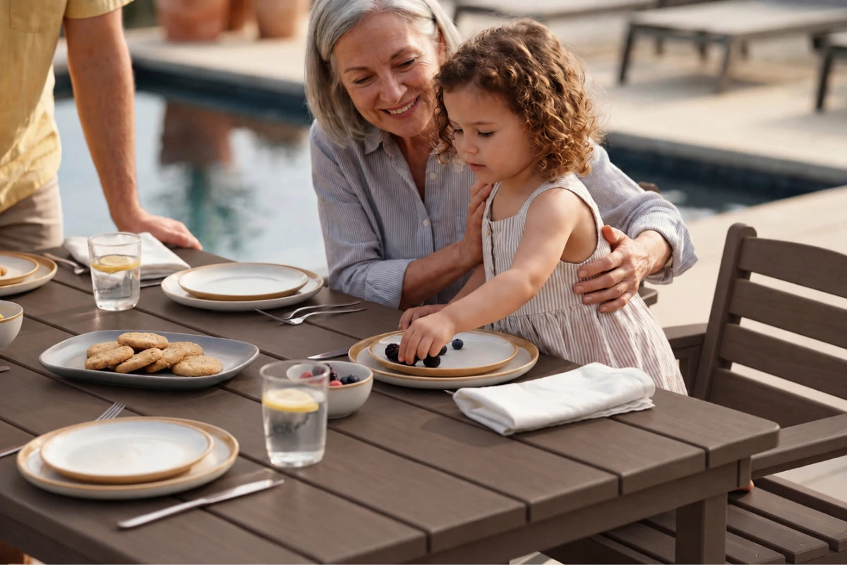 A grandmother and young daughter share a meal at an outdoor coffee-colored dining table by a swimming pool. The table is set with plates, glasses, and a plate of cookies.