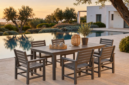 A 7-piece outdoor dining set made of weathered gray wood, including a rectangular table and six matching armchairs, arranged on a stone patio next to a swimming pool at sunset.
