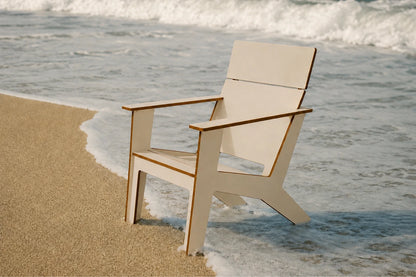 A modern poly Adirondack chair with a beige finish and medium brown edges, positioned on a sandy beach right at the edge of the ocean with waves crashing behind it.