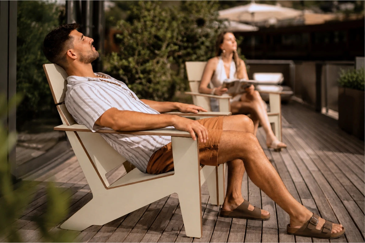 A man relaxes in a modern Adirondack-style outdoor chair on a wooden deck, wearing a striped shirt and brown sandals. A woman sits in a matching chair in the background on a patio.