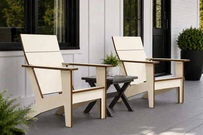 Two modern Adirondack-style outdoor chairs with beige fabric and natural wood frames, paired with a small dark side table on a gray deck in front of a white house with black-framed windows.