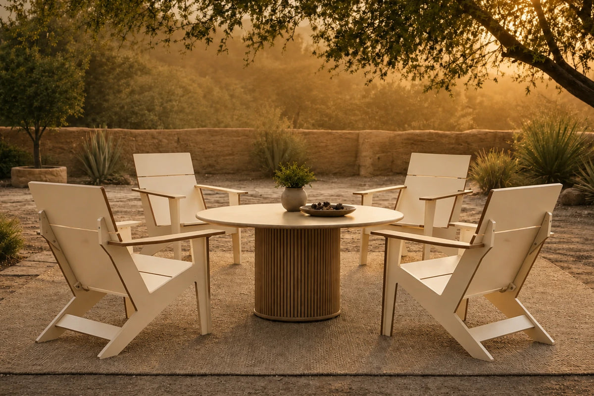 A set of four beige outdoor Adirondack-style chairs arranged around a round slatted pedestal table on a stone patio, with a warm golden sunset background and trees.