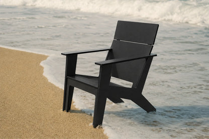 A black modern poly Adirondack chair positioned on a sandy shore with ocean waves washing around it.