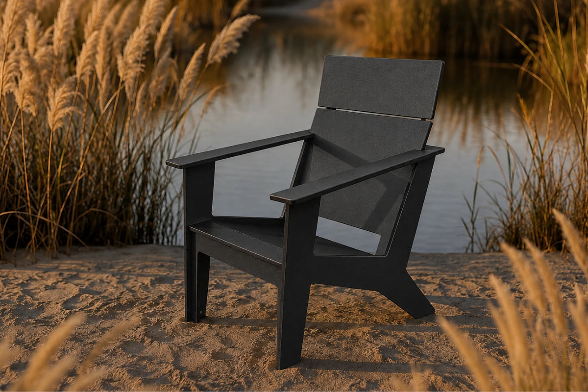 A black outdoor Adirondack chair positioned on a sandy bank next to a calm body of water, framed by golden reeds under warm sunset light.