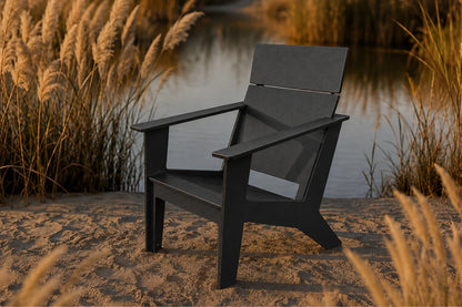 A black outdoor Adirondack chair positioned on a sandy bank next to a calm body of water, framed by golden reeds under warm sunset light.