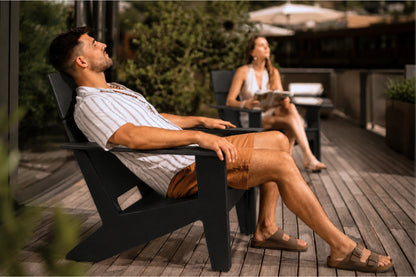 A man relaxes in a black outdoor Adirondack chair on a wooden deck, wearing a white striped shirt, brown shorts, and sandals. A woman sits in a matching chair in the background on a patio.