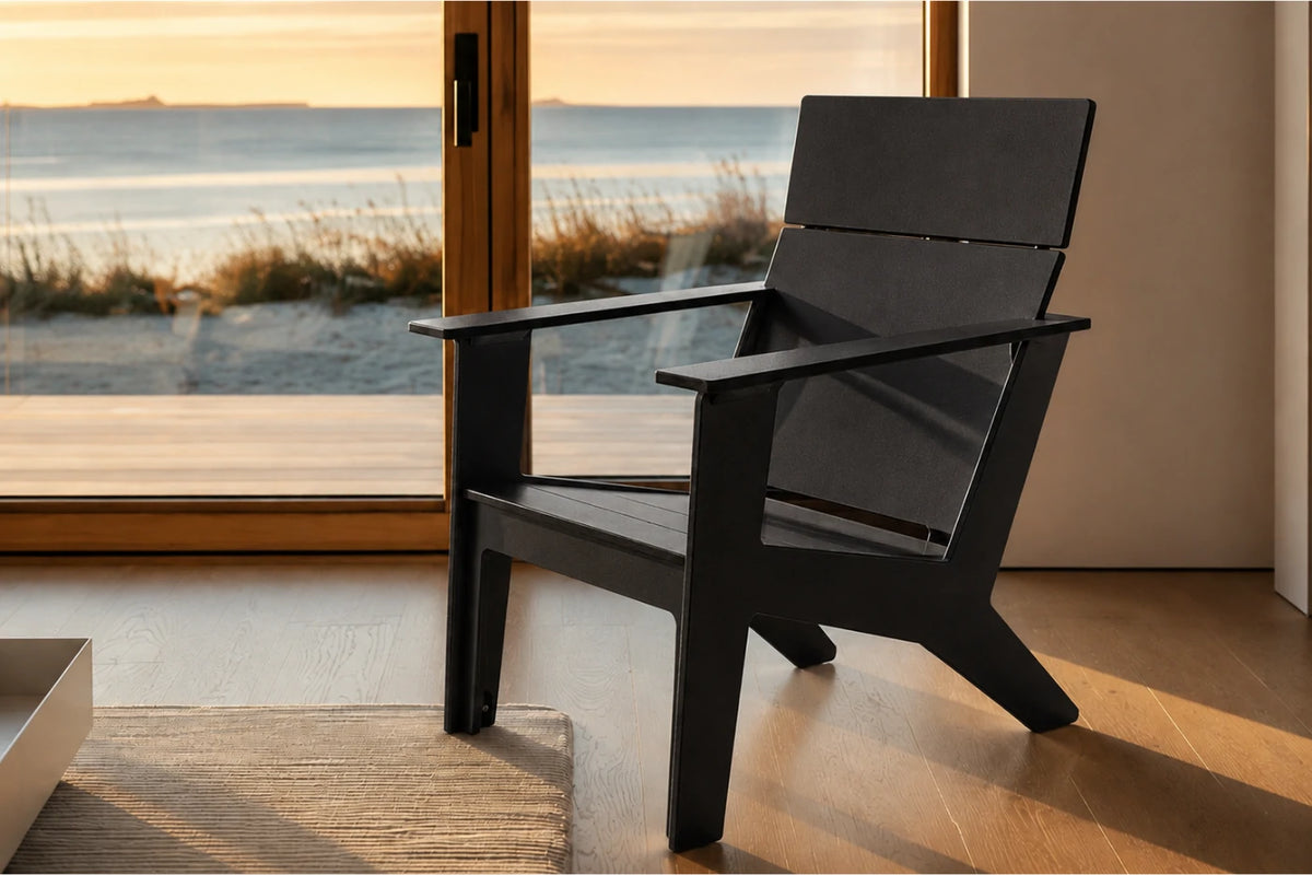A modern black chair with a wide armrest and angled legs, positioned in front of a large glass door overlooking a beach and ocean at sunset.