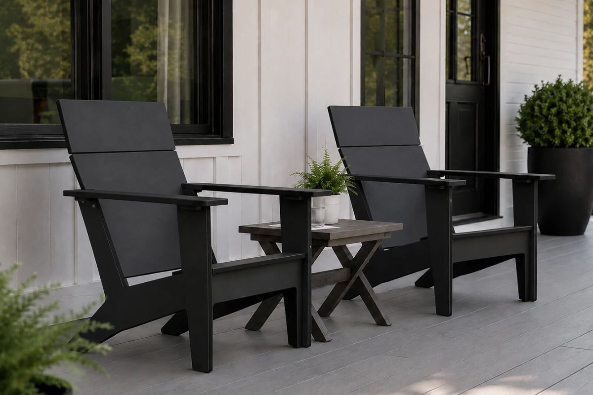 Two black outdoor Adirondack chairs with a small side table between them, set on a gray deck in front of a white house with black-framed windows.