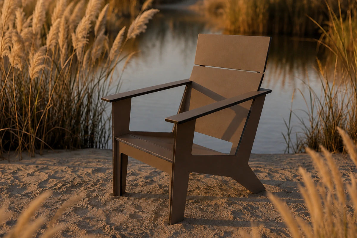 A modern outdoor chair with a geometric, angular design, positioned on a sandy shore next to a calm body of water with tall reeds in the background.