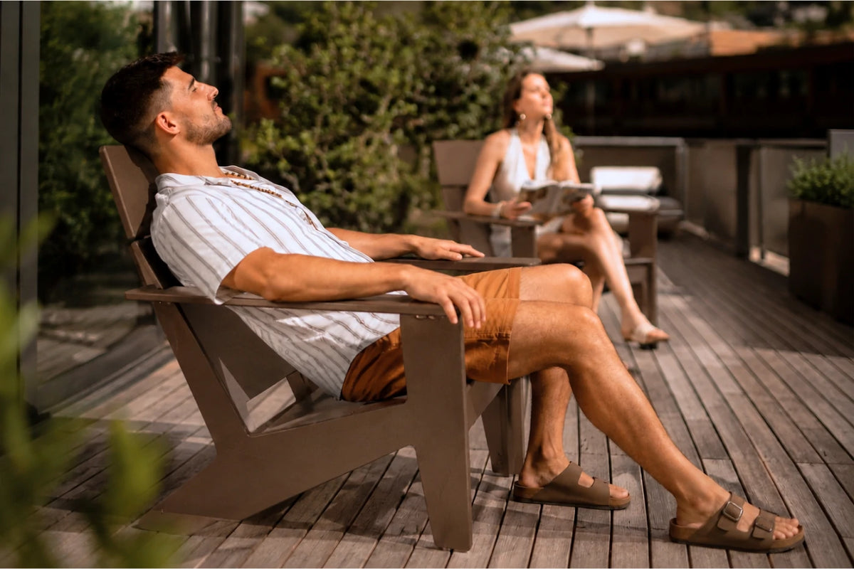 A man relaxes in a coffee modern poly Adirondack chair on a wooden deck, wearing a white striped shirt and brown shorts. A woman sits in a matching chair in the background.