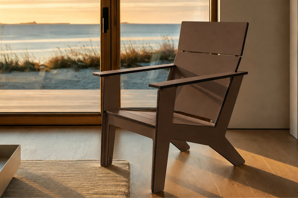 A modern outdoor chair with a wide backrest and armrests, positioned in front of large glass doors looking out onto a beach at sunset.