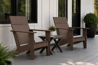 Two coffee outdoor Adirondack chairs with a small matching side table on a light gray porch in front of a white house with black-framed windows.