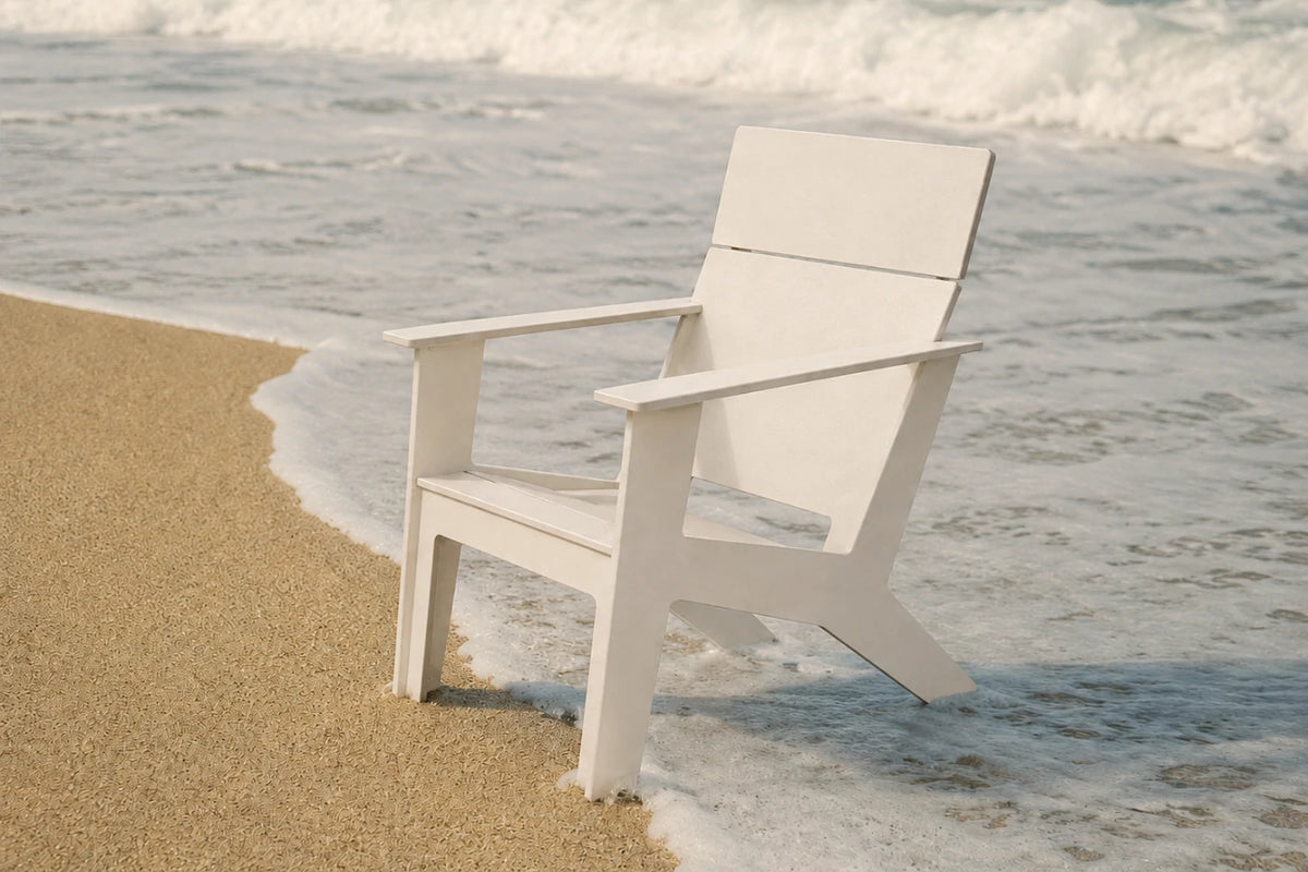 A white modern poly Adirondack chair placed on a sandy shore with ocean waves washing around it.