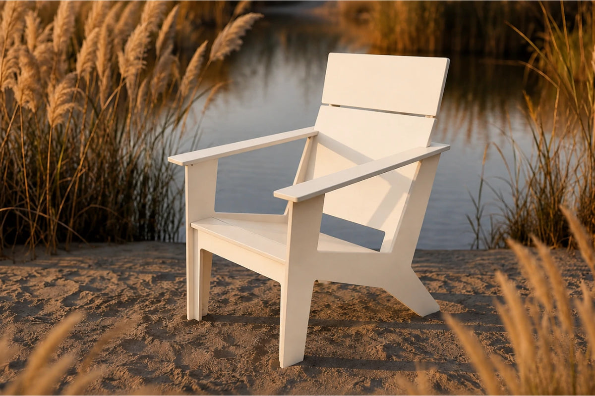 A white outdoor Adirondack-style chair placed on sandy ground beside a calm body of water, with tall golden reeds in the background.