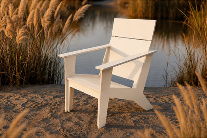A white outdoor Adirondack-style chair placed on sandy ground beside a calm body of water, with tall golden reeds in the background.
