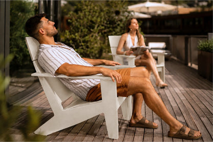 A man relaxes in a white outdoor Adirondack chair on a wooden patio, wearing a striped shirt, brown shorts, and sandals. A woman sits in a matching chair in the background.