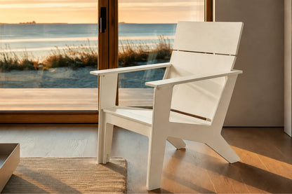 A modern white outdoor Adirondack chair positioned on a wooden deck in front of large glass doors overlooking a beach at sunset.