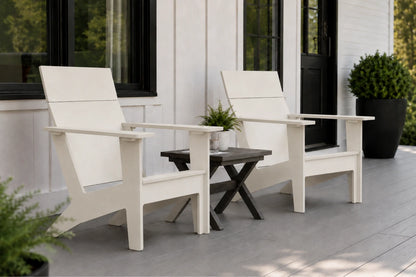 Two white outdoor Adirondack chairs with a small dark wood side table between them, set on a gray porch in front of a white house with black-framed windows.