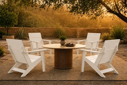Four white outdoor Adirondack-style chairs arranged around a round wooden table on a patio, with trees and warm golden sunlight in the background.