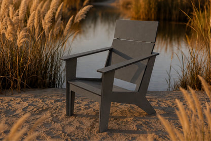 A dark gray modern outdoor Adirondack chair placed on sandy ground next to a calm lake, surrounded by golden reeds at sunset.