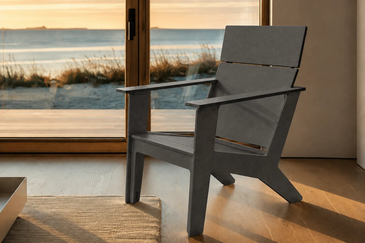 A modern dark gray outdoor Adirondack chair positioned in front of a large glass door overlooking a beach at sunset, with warm golden light reflecting off the wooden floor.