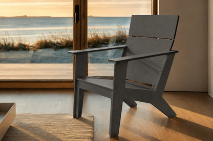 A modern dark gray outdoor Adirondack chair positioned in front of a large glass door overlooking a beach at sunset, with warm golden light reflecting off the wooden floor.