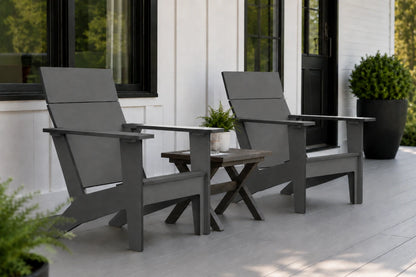 Two dark gray outdoor Adirondack chairs with a small wooden side table between them, set on a light porch in front of a white house with black-framed windows.