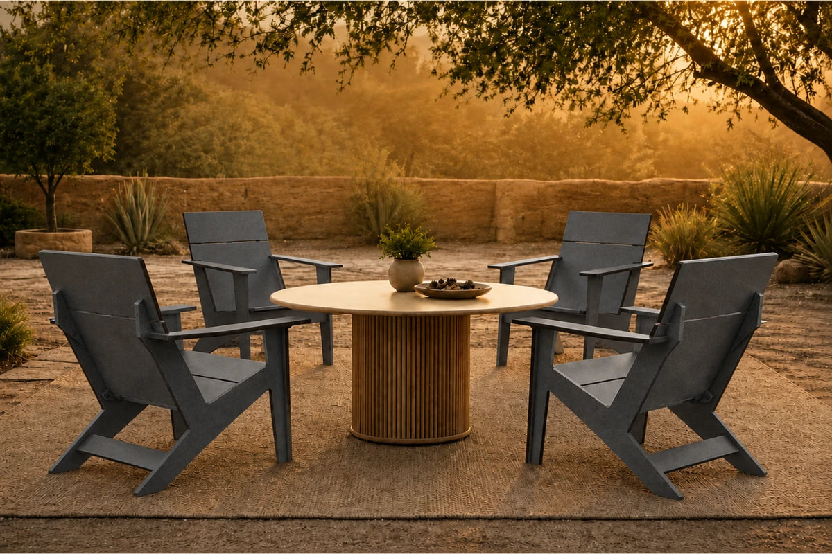 Four dark gray outdoor Adirondack-style chairs arranged around a round pedestal table in a patio setting at sunset, with trees, plants, and a stone wall in the background.
