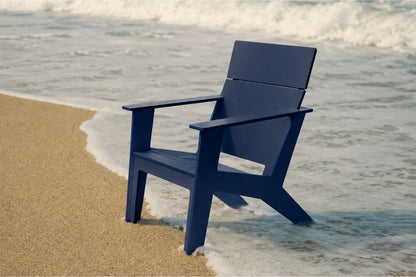 A navy blue Adirondack-style chair placed on a sandy shore with ocean waves washing around it.