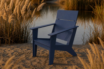 A navy blue Adirondack-style chair placed outdoors on sandy ground beside a calm body of water, with tall golden reeds in the background.
