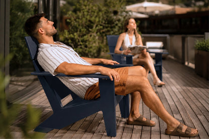 A man relaxes in a navy blue outdoor Adirondack chair on a wooden deck, wearing a white striped shirt and brown shorts. A woman sits in a matching chair in the background.