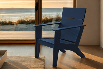 A modern navy blue outdoor chair with a wide backrest and armrests, positioned on a wooden deck in front of large glass doors overlooking a beach at sunset.