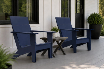 Two navy blue outdoor Adirondack chairs with a small wooden side table between them, set on a light gray porch in front of a white house with black-framed windows.