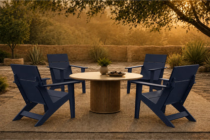 A set of four navy blue outdoor Adirondack-style chairs arranged around a round light wood dining table on a patio, with trees and warm golden evening light in the background.