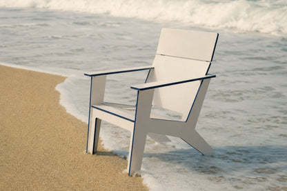A modern white outdoor chair with dark blue trim sits on a sandy beach with ocean waves in the background.
